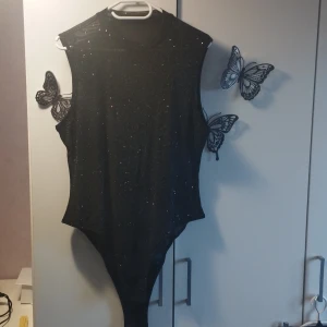 Svart glittrig bodysuit - Snygg svart bodysuit med glittrande detaljer. Den är ärmlös och har en rund halsringning. Perfekt för en kväll ute eller festliga tillfällen. Knäppning i grenen för enkel på- och avtagning.
