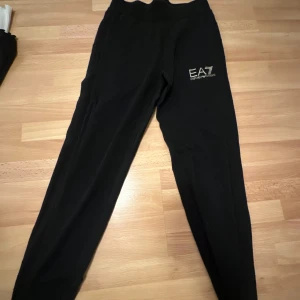Svarta mjukisbyxor från EA7 - Säljer ett par svarta mjukisbyxor från EA7 med logga på sidan. Byxorna har en bekväm passform och är perfekta för en avslappnad stil. De är tillverkade i ett mjukt material som gör dem sköna att bära.