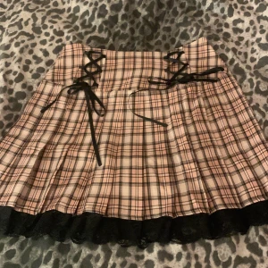 Rutig kjol med snörning och spetsdetalj - Snygg rutig kjol i rosa och svart med snörning framtill och spetskant nedtill. Perfekt för en trendig look!