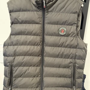 Mörkgrön dunväst från Giorgio Di Mare -Medium  - Snygg mörkgrön dunväst från Giorgio Di Mare med dragkedja och ett stilrent märke på bröstet. Perfekt för kyligare dagar och ger en sportig look. Västen har praktiska fickor med dragkedja.