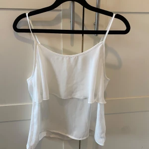Vit linnetopp med volang - Säljer en söt vit linnetopp med tunna axelband och volangdetalj framtill. Perfekt för varma sommardagar eller som ett lager under en kavaj. Toppen är luftig och lätt att matcha med olika stilar.