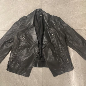 Svart skinnjacka från 157 - Snygg svart skinnjacka från 157 med dragkedjor och knappar. Jackan har en klassisk bikerstil med flera fickor och detaljer som ger en cool look. Tecken på slitage då den är vintage.