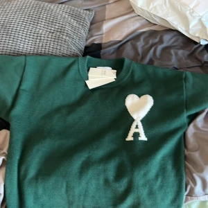 Grön tröja från Ami - Säljer en snygg grön tröja från Ami med ett vitt hjärta och bokstaven A på bröstet. Tröjan har en klassisk rund halsringning och är perfekt för en stilren look. Passar bra till både jeans och chinos. Perfekt för dig som vill ha något unikt i garderoben! Såklart äkta och kan gå ner nån 100-200 för snabb affär