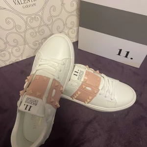 Valentino Rockstud sneakers  - Snygga Valentino Rockstud sneakers i vitt med rosa detaljer. Skorna har de ikoniska nitarna på sidorna och snörning framtill. Perfekta för att ge din outfit en lyxig touch, nya