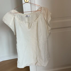 Vit blus från co'couture - Säljer en söt vit blus från co'couture med korta ärmar och en smockad halsringning. Blusen har en avslappnad passform och är perfekt för varma dagar. Den är tillverkad i ett lätt och luftigt material. 