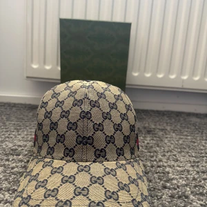 Beige Gucci keps - Snygg beige keps från Gucci med det ikoniska monogrammönstret i svart. Kepsen har en röd och blå randdetalj på sidan och justerbar rem baktill för perfekt passform. Perfekt för att ge din outfit en lyxig touch.