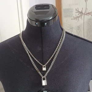 Silverfärgat halsband med hänglås - Dubbelkedjat halsband i silverfärg med två hänglås som detaljer. Kedjorna har olika längd för en lager-på-lager-effekt. Perfekt för att ge en edgy touch till din outfit. Det är två olika halsband men kommer som ett par.