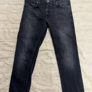 Nudie Jeans Svarta - Snygga svarta jeans från Nudie Jeans med klassisk femficksdesign och knappgylf. De har en diskret tvättad look och märkets signaturbroderi på bakfickorna. Perfekta för en stilren och avslappnad look. Size 32/32