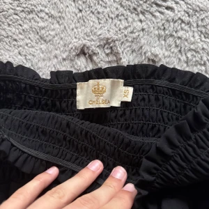Svart volangkjol från Chelsea XS - Supersöt svart kortkjol från Chelsea med bred smock i midjan och dubbla volanger. Köpt i Chelsea affär och används några par gånger. Säljer pågrund av att den inte kommer mer till användning.