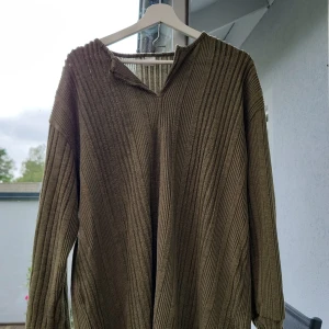 Olivgrön ribbad tröja 1XL - Olivgrön långärmad tröja i ribbat material med avslappnad passform. Tröjan har en liten v-ringning vid halsen och är tillverkad i mjuk polyesterblandning. Perfekt för lager-på-lager under kyliga dagar.