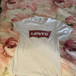 Vit Levi's t-shirt med röd logga XS - Vit t-shirt från Levi's i bomull med klassisk röd logga framtill. Modellen är kortärmad och har en rund halsringning. Perfekt basic-plagg med snygg streetkänsla och ikonisk branding.
