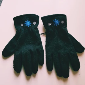 Svarta fleecehandskar med snöflingor strl 7 - Mysiga svarta fleecehandskar med broderade snöflingor i blått och vitt vid handleden. Perfekta för kalla vinterdagar och har en mjuk, fluffig känsla. Femfingermodell med elastisk handled för bättre passform.  Helt skick dock noppror
