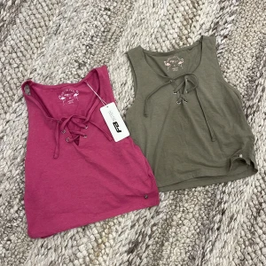 Två croppade toppar med snörning - Säljer två croppade toppar en i mörkrosa och en i olivgrön. Båda har snygg snörning framtill och är ärmlösa. Säljer båda tillsammans. En är helt ny och har fortfarande prislappen på. 20 kr tillsammans 