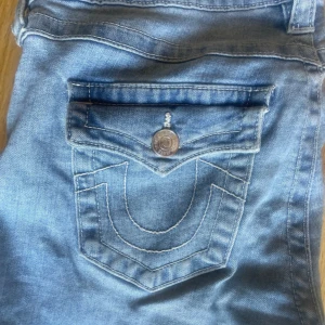 True Religion Joey ljusblå flare jeans - Säljer ett par ikoniska True Religion Joey jeans i ljusblå tvätt med snygga kontrastsömmar och stora fickor med lock bak. Modellen är low rise med rejäl flare och bootcut, perfekt för dig som gillar retrovibbar. Klassisk True Religion patch och detaljerade knappar.