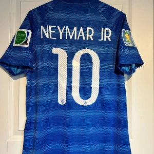 Blå Brasilien Neymar JR VM 2014 tröja - Säljer en blå Brasilien fotbollströja från VM 2014 med Neymar JR och nummer 10 på ryggen. Tröjan har korta ärmar CBF-emblem och FIFA World Cup-patchar på båda ärmarna. Tillverkad i lätt och ventilerande Dri-FIT-material.