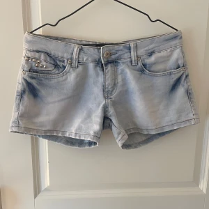 Ljusblå jeansshorts med nitar D&R - Snygga ljusblå jeansshorts från Dolce & Rosa med coola silvriga nitar på fickan. Klassisk femficksmodell, låg midja och slitna detaljer. Perfekta för sommarens alla äventyr. Styla med en oversized tee eller crop top för en chill vibe.