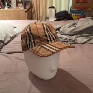 Burberry rutig keps i beige och brunt - Snygg keps från Burberry i klassiskt rutigt mönster med beige, bruna, svarta och röda toner. Justerbar rem baktill i läder och böjd skärm. Perfekt accessoar för en trendig streetstyle-look. Riktig fin keps sjukt bra skick