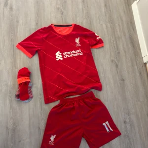 Liverpool Salah matchställ röd - Liverpool FC matchställ med Salah 11 på ryggen. Tröjan är röd med orange detaljer, kortärmad och har vita tryck med klubbmärke och sponsor. Tillhörande shorts och strumpor i rött och orange. Materialet är lätt och andas, perfekt för fotbollsträning eller match.