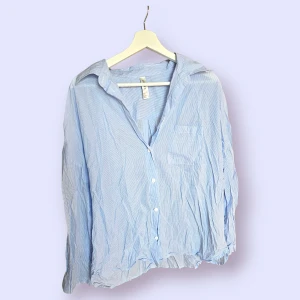 Randig skjorta i storlek L från Lindex - Ljusblå skjorta med vita ränder och långa ärmar. Skjortan har en bröstficka och knäpps med knappar. Aldrig använd.