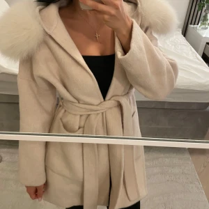 Beige kappa med avtagbar pälskrage Borninstockholm - Säljer en beige kappa från Born In Stockholm med stor huva och fluffiga pälskragar på axlarna. Jackan har bälte i midjan för snygg passform och är tillverkad i mjukt material. 