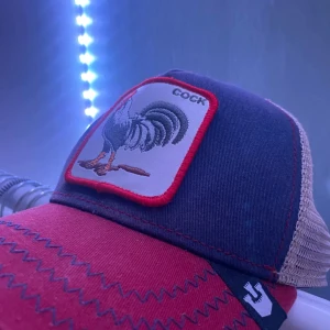 Röd och blå truckerkeps från The Farm - Säljer en snygg truckerkeps från The Farm med röd skärm, blå front och beige mesh baktill. Framtill finns ett broderat tuppmärke med röd kant och texten 'COCK'. Kepsen har coola zigzag-sömmar och justerbar passform.