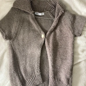 Beige stickad väst från Zara - Mysig beige stickad väst från Zara med korta ärmar och stor krage. Västen har en enkel knapp framtill och ribbade detaljer vid krage och nederkant. Perfekt att slänga över en långärmad tröja för extra värme.