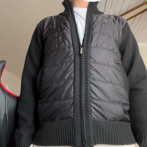 Svart stickad pufferjacka med dragkedja - Säljer en svart jacka med stickade ärmar och rygg samt quiltad pufferfront. Jackan har ribbade muddar, hög krage och dragkedja framtill. Perfekt för dig som vill ha en chill och stilren look med lite extra edge. Skick 8/10 storlek S men passar också M. Hör av dig om de är något du undrar över☺️ Priset ej hugget i sten🤩