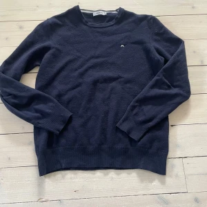 Mörkblå stickad tröja J.Lindeberg XL - Mörkblå stickad tröja från J.Lindeberg i storlek XL. Klassisk rund halsringning och diskret logga broderad på bröstet. Tröjan har långa ärmar och ribbade muddar för en snygg och enkel look. Perfekt för lager-på-lager under kyliga dagar. Den har ett litet hål på armbågen som knappt syns och som lätt går att sy igen 