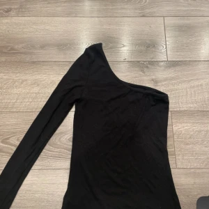 Svart oneshoulder topp - Snygg svart topp med oneshoulder-design och lång ärm. Enkel och stilren modell som passar perfekt till jeans eller kjol. Mjuk och stretchig känsla i materialet för en bekväm passform.