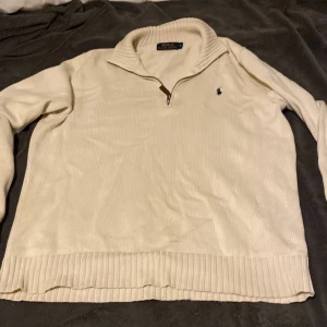 Beige stickad tröja Polo Ralph Lauren - Beige stickad tröja från Polo Ralph Lauren med halv dragkedja och ribbad krage. Klassisk broderad logga på bröstet och ribbade muddar. Perfekt för lager-på-lager och chill stil under höst och vinter.