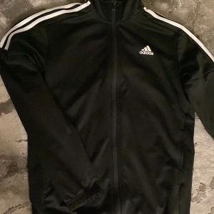Svart Adidas kofta med vita ränder - Svart kofta från Adidas med klassiska vita ränder längs ärmarna och vit Adidas-logga på bröstet. Mycket bra skick!