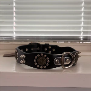 Svart skärp med nitar och BACKBONE-tag - Säljer ett svart hundhalsband i äkta skinn från BACKBONE. Halsbandet har coola silverfärgade nitar, stora öglor och detaljerade metallplattor med mönster. Spännet är robust i metall. Helt nytt, lappen sitter kvar. 60CM NYPRIS 705kr