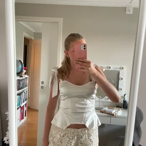 Vit satin korsett med knytband - Supersnygg vit korsettopp i glansig satin med breda axelband som knyts i rosetter. Figurnära passform med markerade sömmar och lätt utsvängd nederkant. Perfekt för att lyfta vilken outfit som helst!