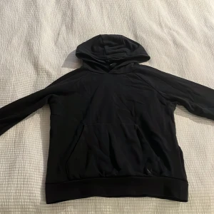 Svart hoodie från SOC - Svart hoodie från SOC med klassisk huva och magficka. Tröjan har lång ärm och är tillverkad i mjuk bomullsmix som är skön att ha på sig. Perfekt för chill dagar eller när du vill ha en enkel och stilren look.