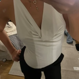 Vit omlott ärmlös topp med öppen rygg - Säljer en vit omlott-topp med djup v-ringning och öppen rygg. Toppen är ärmlös och har en snygg draperad effekt framtill. Perfekt för dig som gillar stilrena och trendiga plagg med en twist.