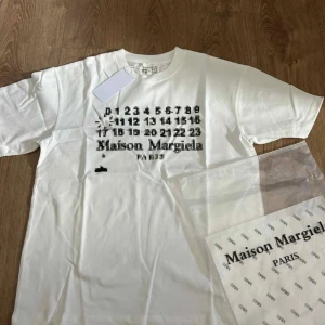 Maison Margiela vit t-shirt med print - Vit t-shirt från Maison Margiela med svart pixel-inspirerat tryck och logga framtill. Klassisk passform med rund hals och korta ärmar. Tillverkad i mjuk bomull för en clean och modern look.
