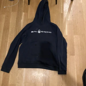 Svart hoodie från Sail Racing - Svart hoodie från Sail Racing med vit logga tryckt på bröstet. Klassisk modell med huva och känguruficka framtill. Perfekt för dig som gillar en sportig och avslappnad stil. Mjuk och skön, passar till jeans eller mjukisbyxor.