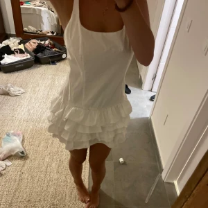 Vit kort volangklänning med axelband - Supersöt vit kort klänning med breda axelband och flera lager volanger nedtill. Den har en rak urringning och sitter snyggt figurnära upptill. Perfekt för sommarens varma dagar och ger en fräsch och lekfull look. och har inbyggda shorts 