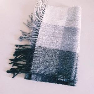 Rutig halsduk i ull från Tippy - Snygg rutig halsduk från Tippy i olika nyanser av grått och svart. Halsduken är lång med fransar i ändarna och är tillverkad i 100% ull, vilket gör den både varm och mjuk. Perfekt accessoar för att lyfta din vinterstil.  Helt skick dock noppror