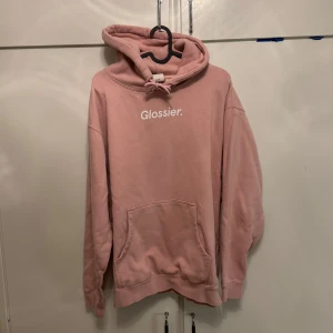 Rosa Glossier hoodie med huva - Säljer en rosa hoodie från Glossier med vit logga framtill. Tröjan har huva med snörning och en stor magficka. Knappt använd. 