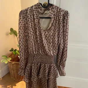 Mönstrad kortklänning Sofie Schnoor - Supersöt kortklänning från Sofie Schnoor med volangkjol, smock i midjan och långa ärmar. Klänningen har ett ljust, blommigt mönster i beige och bruna toner samt knytband vid halsen. Perfekt för dig som gillar romantiska och feminina detaljer.