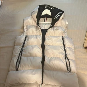 Dunväst från Moncler med huva - Snygg beige/grå dunväst från Moncler med svart huva och dragkedja framtill. Helt ny! Strl s passar både dam och herr.