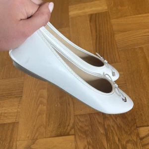 ballerinaskor - Snygga vita ballerinaskor med rund tå och dekorativ rosett framtill. Skorna har en platt sula och är tillverkade i ett slätt syntetmaterial som ger en clean och fräsch look. Perfekta för dig som gillar en stilren och klassisk vibe.