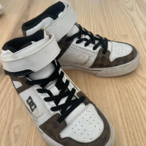 DC Shoes rutiga high top sneakers - Snygga high top sneakers från DC Shoes i vitt och brunt med rutigt mönster och guldiga DC-loggor. Skorna har svart foder, svarta skosnören och bred kardborre över vristen. Ovandelen är i skinn och syntet, perfekt för dig som gillar streetstyle.