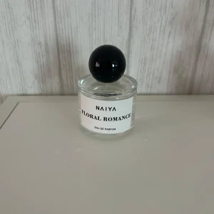 NAIYA Floral Romance parfym - NAIYA Floral Romance Eau de Parfum i en stilren, rund glasflaska med svart klotformad kork. Flaskan är transparent med vit etikett och svart text. Perfekt för dig som gillar blommiga dofter och snygg minimalistisk design.