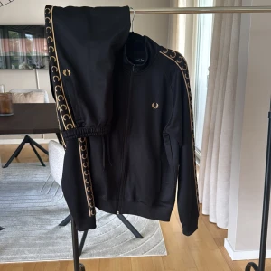 Svart track suit från Fred Perry - Svart track suit från Fred Perry med guldiga detaljer och logga längs ärmarna. Jackan har dragkedja framtill, ribbade muddar och klassisk sportig look. Perfekt för dig som gillar streetwear och vill sticka ut med snygga kontraster.