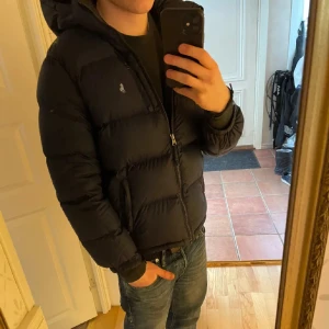 Svart dunjacka från Ralph Lauren - Säljer en svart dunjacka från Ralph Lauren med huva och dragkedja. Jackan är quiltad och har en klassisk pufferstil med diskret logga på bröstet. Perfekt för kalla dagar och riktigt snygg till jeans.