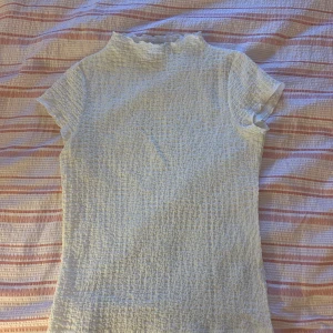 Vit smockad topp från Kappahl - Supersöt vit topp från Kappahl i smockat tyg med korta ärmar och liten volangkant vid halsen. Toppen har en tight passform och är perfekt för dig som gillar en feminin och stilren look. Enkel att matcha med både jeans och kjol.