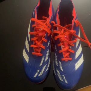 Adidas Predator blå fotbollsskor - Säljer ett par Adidas Predator fotbollsskor i blått med vita detaljer och röda snören. Skorna har klassisk design med tydliga ränder på sidorna och är tillverkade i syntetmaterial för bästa grepp på planen. Perfekta för dig som vill sticka ut på matchen!