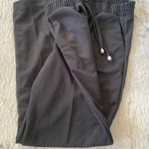 Svarta mjukisbyxor med snörning - Svarta mjukisbyxor med elastisk midja och snörning med silverfärgade detaljer. Byxorna har raka ben och är tillverkade i ett mjukt material som känns skönt mot huden. Perfekta för chill dagar eller när du vill vara bekväm men ändå se stilren ut.                         Den är i storlek L men är som storlek S/M